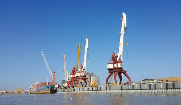 Large port cranes stand tall by the waterfront, with a cargo ship docked nearby. The sky is a clear blue, setting a contrasting backdrop to the industrial scene. The surrounding area includes warehouses and storage facilities, while seagulls hover above the water.