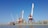 Large port cranes stand tall by the waterfront, with a cargo ship docked nearby. The sky is a clear blue, setting a contrasting backdrop to the industrial scene. The surrounding area includes warehouses and storage facilities, while seagulls hover above the water.