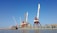 Large port cranes stand tall by the waterfront, with a cargo ship docked nearby. The sky is a clear blue, setting a contrasting backdrop to the industrial scene. The surrounding area includes warehouses and storage facilities, while seagulls hover above the water.
