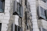 A close-up of a modern building facade featuring reflective, metallic panels with an undulating design. Several windows with dark frames protrude from the structure, adding to the building's dynamic appearance.
