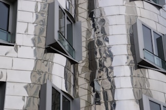 A close-up of a modern building facade featuring reflective, metallic panels with an undulating design. Several windows with dark frames protrude from the structure, adding to the building's dynamic appearance.