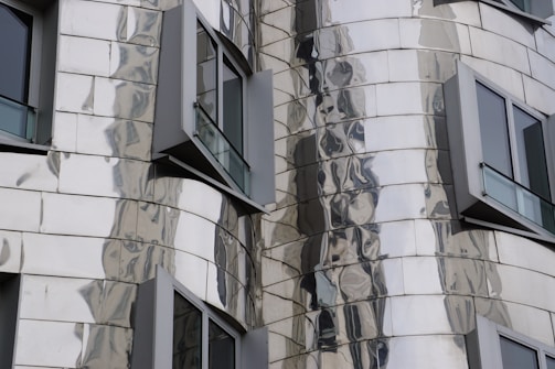 A close-up of a modern building facade featuring reflective, metallic panels with an undulating design. Several windows with dark frames protrude from the structure, adding to the building's dynamic appearance.