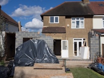 A two-story house under renovation with new brickwork being added. A black tarp covers construction materials in the foreground, surrounded by wood and other building materials. The house features a light brown exterior with two windows on the upper floor, white-framed doors, and a partially completed extension on the ground floor.