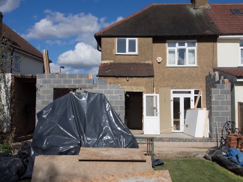 A two-story house under renovation with new brickwork being added. A black tarp covers construction materials in the foreground, surrounded by wood and other building materials. The house features a light brown exterior with two windows on the upper floor, white-framed doors, and a partially completed extension on the ground floor.