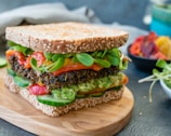 A healthy sandwich on a wooden serving board, made with seeded whole grain bread filled with layers of fresh vegetables including slices of cucumber, leafy greens, roasted red peppers, guacamole, and a black bean patty.