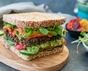 A healthy sandwich on a wooden serving board, made with seeded whole grain bread filled with layers of fresh vegetables including slices of cucumber, leafy greens, roasted red peppers, guacamole, and a black bean patty.