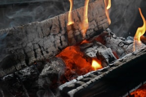 Burning logs with charred surfaces are engulfed in bright yellow flames. The wood glows with orange embers, and gray smoke rises, creating an atmospheric and warm scene.