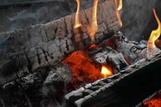 Burning logs with charred surfaces are engulfed in bright yellow flames. The wood glows with orange embers, and gray smoke rises, creating an atmospheric and warm scene.