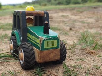 A wooden toy tractor sits on a patch of dirt and grass. The tractor is painted green with black wheels and has a driver inside wearing a yellow helmet. The word 'Tractor' is written on the side. The surroundings are blurred out with green foliage in the background.
