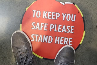 A pair of gray sneakers standing on a circular red floor sign with the text 'TO KEEP YOU SAFE PLEASE STAND HERE'. The sign is bordered with yellow and black and is positioned on a gray concrete floor.