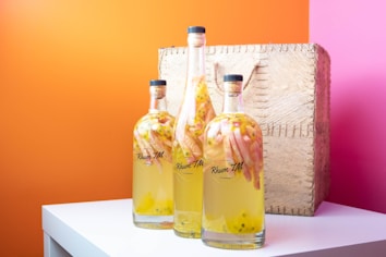 Three bottles of infused rum with fruit inside are arranged on a white table. The bottles are of different sizes and have cork stoppers. They are set against a vibrant background with orange and pink walls. A textured wicker basket is positioned behind the bottles, adding a rustic element.