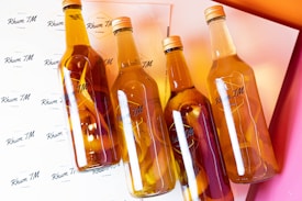 Four clear glass bottles filled with a golden liquid and citrus peels are arranged diagonally on a background covered with the label 'Rhum TM'. The light reflects off the bottles and there are gradients of orange and pink in the background, creating a warm and vibrant atmosphere.