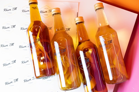 Four clear glass bottles filled with a golden liquid and citrus peels are arranged diagonally on a background covered with the label 'Rhum TM'. The light reflects off the bottles and there are gradients of orange and pink in the background, creating a warm and vibrant atmosphere.
