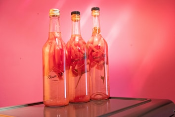 Three bottles filled with a pink liquid, each containing visible slices of fruit. Positioned against a vibrant pink background, the bottles are labeled with the text 'Rhum TM' and rest atop a reflective surface.