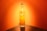 A tall, clear glass bottle filled with a yellow liquid is placed against a bright orange background. The bottle's label reads 'Rhum JM' and it is positioned on a reflective surface that mirrors part of the bottle and the vibrant color of the background.