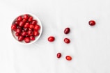 A white bowl filled with bright red cranberries is placed on a white surface. Several cranberries are scattered around the bowl, adding a casual, natural aesthetic.