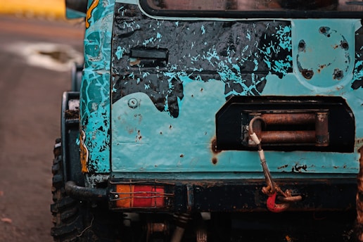 The image features the rear section of a rugged off-road vehicle with a distressed blue and black paint job. There is visible rust and wear on the metal surface, and a red tow hook is attached to a winch on the back.
