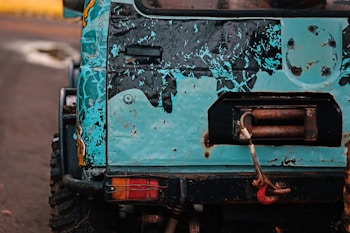 The image features the rear section of a rugged off-road vehicle with a distressed blue and black paint job. There is visible rust and wear on the metal surface, and a red tow hook is attached to a winch on the back.