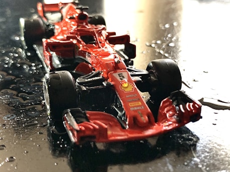 A miniature red Formula 1 race car on a wet, reflective surface. The car features detailed branding and design elements, including sponsor logos, with water droplets scattered around.