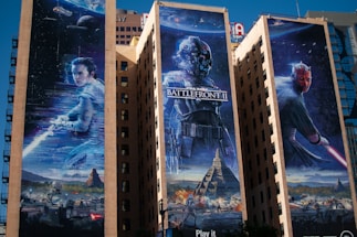 Three large murals on the sides of tall buildings depict scenes from a popular sci-fi franchise. The left mural features a character holding a glowing weapon against a cosmic backdrop, the middle showcases a masked figure in futuristic armor with the text 'Battlefront II,' and the right displays a red-faced antagonist wielding a similar glowing weapon. Below, a scene of battle with spaceships and rugged terrain is visible.