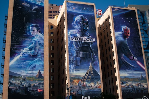 Three large murals on the sides of tall buildings depict scenes from a popular sci-fi franchise. The left mural features a character holding a glowing weapon against a cosmic backdrop, the middle showcases a masked figure in futuristic armor with the text 'Battlefront II,' and the right displays a red-faced antagonist wielding a similar glowing weapon. Below, a scene of battle with spaceships and rugged terrain is visible.