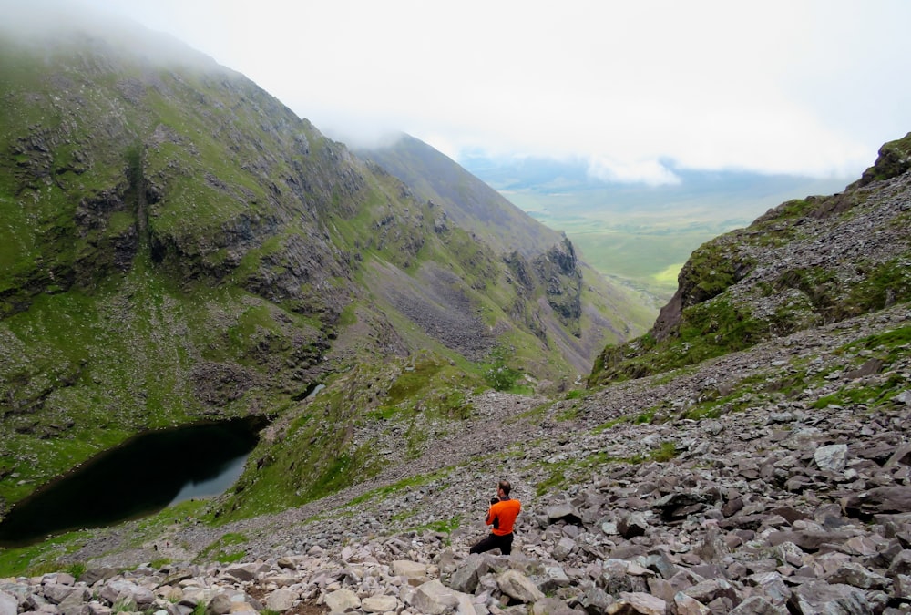 person in orange jacket sitting on rocky ground near green mountains during daytime