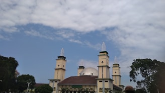 A large mosque with multiple tall minarets and a prominent dome. The building is surrounded by lush greenery and trees, set against a partly cloudy sky.