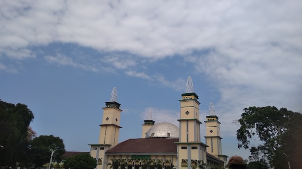 A large mosque with multiple tall minarets and a prominent dome. The building is surrounded by lush greenery and trees, set against a partly cloudy sky.