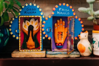 Two decorated shrines featuring iconic elements: one with Diego Rivera's name above a floral depiction, the other with Frida Kahlo's name and an image of her within a hand sculpture. Surrounding the shrines are decorative plants and a ceramic bird.
