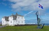 A traditional white house with a steep dark roof sits near the shoreline. In front of the house is a statue of a figure beside a metal structure resembling a sail or harp. A flagpole displaying the Icelandic flag stands nearby, waving against a backdrop of a partly cloudy blue sky.