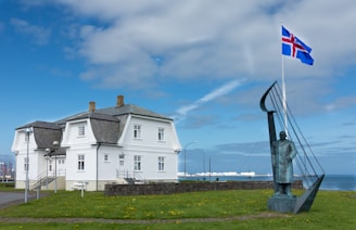 A traditional white house with a steep dark roof sits near the shoreline. In front of the house is a statue of a figure beside a metal structure resembling a sail or harp. A flagpole displaying the Icelandic flag stands nearby, waving against a backdrop of a partly cloudy blue sky.