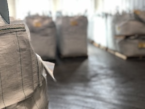 Large industrial sacks are positioned in a warehouse with a focus on one sack in the foreground and more sacks blurred in the background. The setting appears to be indoors with soft lighting, creating a sense of a spacious storage facility.