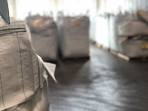 Large industrial sacks are positioned in a warehouse with a focus on one sack in the foreground and more sacks blurred in the background. The setting appears to be indoors with soft lighting, creating a sense of a spacious storage facility.