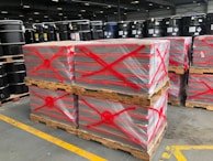 Stacks of pallets wrapped in plastic are marked with red spray-painted crosses. Black industrial barrels are arranged on shelves in the background, and the scene is set in a large industrial warehouse with high ceilings and fluorescent lighting.