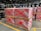 Stacks of pallets wrapped in plastic are marked with red spray-painted crosses. Black industrial barrels are arranged on shelves in the background, and the scene is set in a large industrial warehouse with high ceilings and fluorescent lighting.