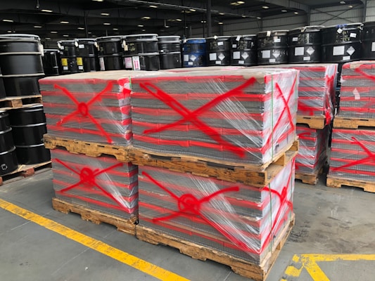 Stacks of pallets wrapped in plastic are marked with red spray-painted crosses. Black industrial barrels are arranged on shelves in the background, and the scene is set in a large industrial warehouse with high ceilings and fluorescent lighting.