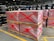 Stacks of pallets wrapped in plastic are marked with red spray-painted crosses. Black industrial barrels are arranged on shelves in the background, and the scene is set in a large industrial warehouse with high ceilings and fluorescent lighting.