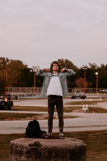 A person stands confidently on a large concrete platform in an open area, adjusting their jacket. The scene includes a backpack resting nearby, a road with moving vehicles in the background, and a park-like setting with scattered trees. The sky is overcast and the setting appears to be late afternoon or early evening.