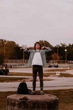 A person stands confidently on a large concrete platform in an open area, adjusting their jacket. The scene includes a backpack resting nearby, a road with moving vehicles in the background, and a park-like setting with scattered trees. The sky is overcast and the setting appears to be late afternoon or early evening.
