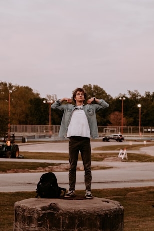 A person stands confidently on a large concrete platform in an open area, adjusting their jacket. The scene includes a backpack resting nearby, a road with moving vehicles in the background, and a park-like setting with scattered trees. The sky is overcast and the setting appears to be late afternoon or early evening.