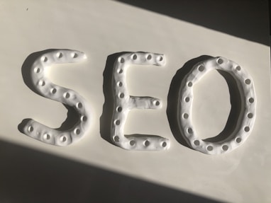 Three-dimensional letters spelling 'SEO' are crafted from a white, dough-like material. Each letter features a series of small circular indentations, creating a textured pattern. The arrangement is placed against a smooth white surface, with a shadow cast diagonally across.