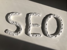 Three-dimensional letters spelling 'SEO' are crafted from a white, dough-like material. Each letter features a series of small circular indentations, creating a textured pattern. The arrangement is placed against a smooth white surface, with a shadow cast diagonally across.