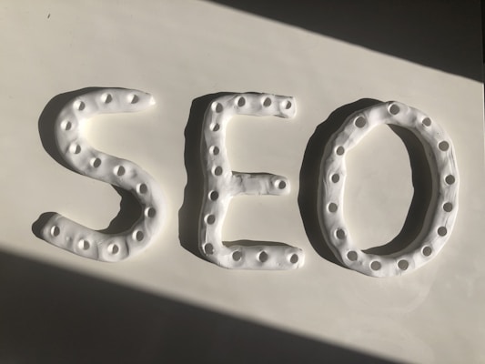 Three-dimensional letters spelling 'SEO' are crafted from a white, dough-like material. Each letter features a series of small circular indentations, creating a textured pattern. The arrangement is placed against a smooth white surface, with a shadow cast diagonally across.