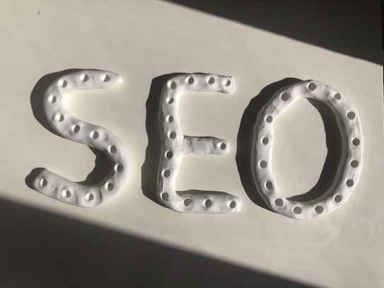 Three-dimensional letters spelling 'SEO' are crafted from a white, dough-like material. Each letter features a series of small circular indentations, creating a textured pattern. The arrangement is placed against a smooth white surface, with a shadow cast diagonally across.