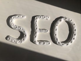 Three-dimensional letters spelling 'SEO' are crafted from a white, dough-like material. Each letter features a series of small circular indentations, creating a textured pattern. The arrangement is placed against a smooth white surface, with a shadow cast diagonally across.