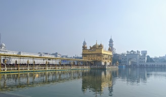 A stunning golden temple stands prominently in the center, with intricate architecture and a large body of water surrounding it, creating a serene reflection. A long bridge filled with people approaches the temple, and several tourists are seen admiring the view.