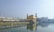 A stunning golden temple stands prominently in the center, with intricate architecture and a large body of water surrounding it, creating a serene reflection. A long bridge filled with people approaches the temple, and several tourists are seen admiring the view.