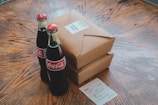 Two glass Coca-Cola bottles are placed on a wooden table next to two brown cardboard food packages. The packages have white labels, one of which reads 'Chicken Salad.' A small paper with food order details is also on the table.