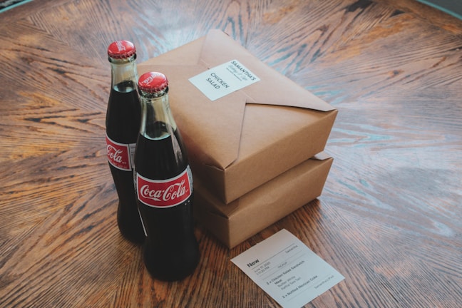 Two glass Coca-Cola bottles are placed on a wooden table next to two brown cardboard food packages. The packages have white labels, one of which reads 'Chicken Salad.' A small paper with food order details is also on the table.