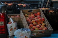 A collection of large crates filled with ripe peaches is placed in the back of a truck. Next to them is a white paper bag decorated with peach illustrations. The surroundings are likely part of a market or farm setting.